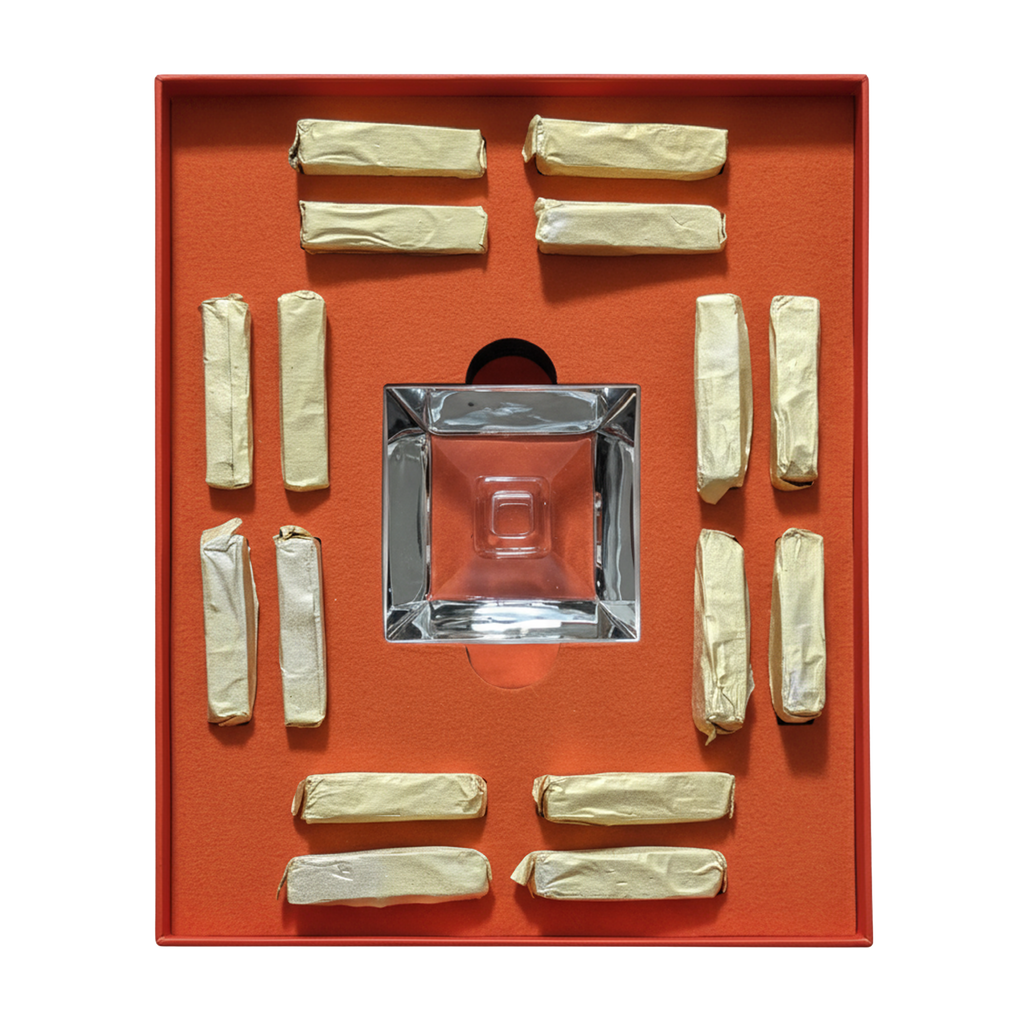 A square clear glass ashtray displayed in a red-orange presentation box, surrounded by multiple cream-colored rolled paper napkins on a white background.