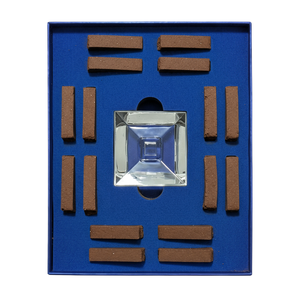 A square blue presentation box containing a clear faceted glass or acrylic pyramid in the center, surrounded by eight small brown rectangular sticks arranged in a grid.