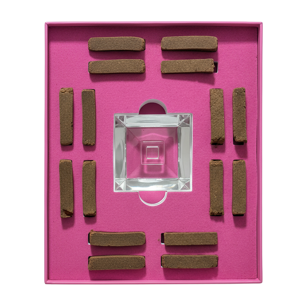 A square pink presentation box containing a clear glass square dish in the center and eight brown rectangular incense sticks arranged around it on a bright pink insert.