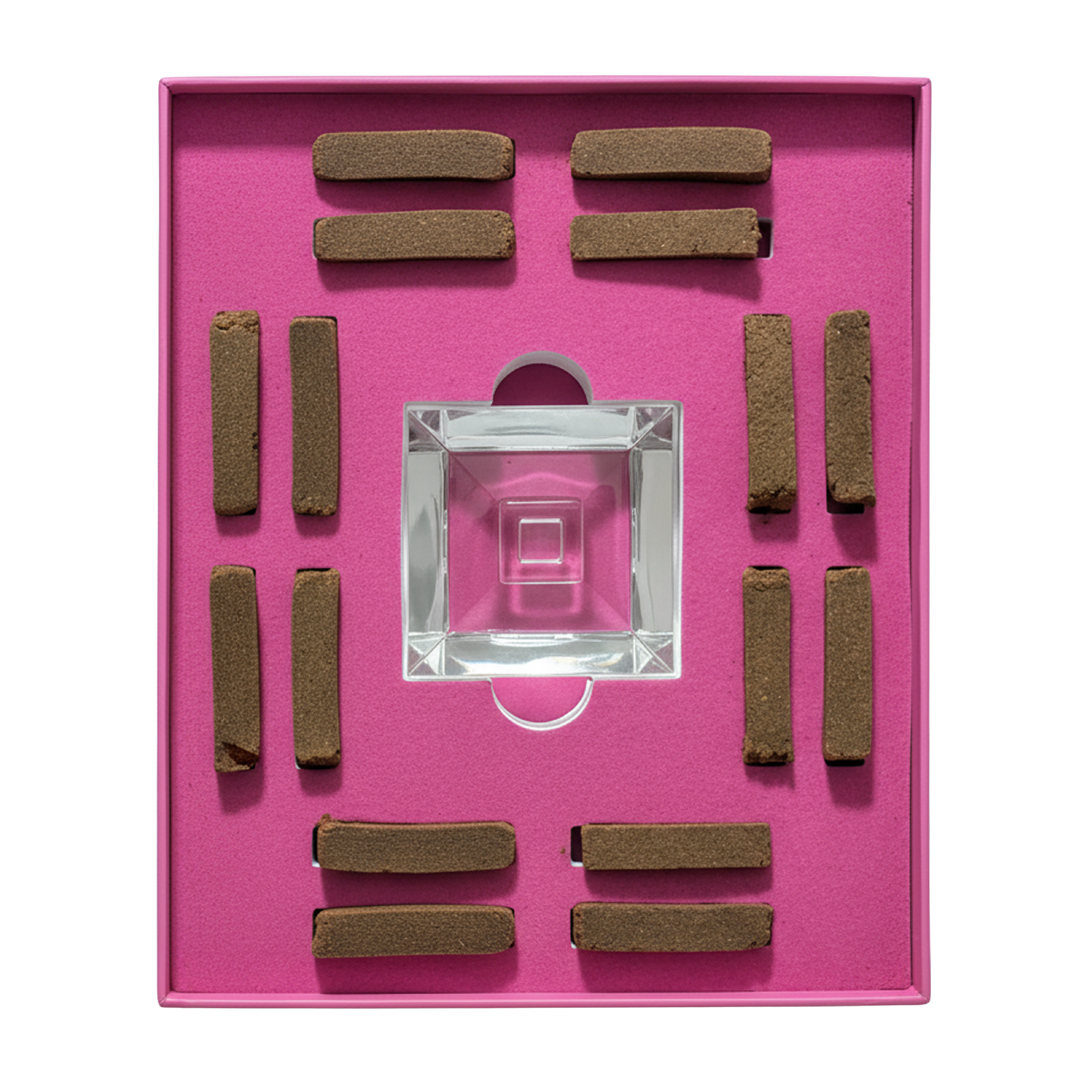 A square pink presentation box containing a clear glass square dish in the center and eight brown rectangular incense sticks arranged around it on a bright pink insert.